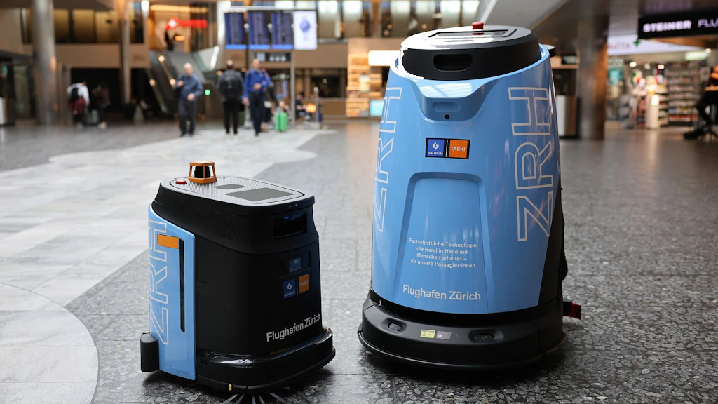 Zwei autonome Reinigungsroboter in einem Flughafenterminal reinigen den Boden; im Hintergrund gehen Reisende vorbei. Die blau-schwarzen Geräte navigieren selbstständig, erkennen Hindernisse und arbeiten im laufenden Betrieb.