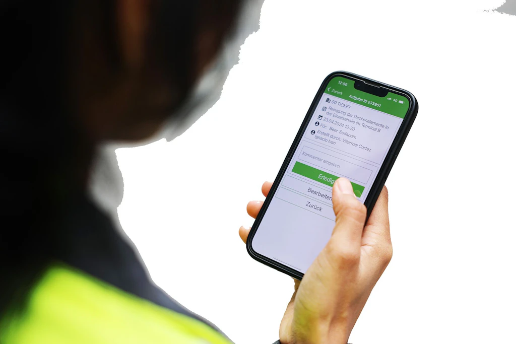 Eine Person hält ein Smartphone und bestätigt in einer App einen Reinigungsauftrag; auf dem Display sind Details zum Ticket zu sehen.
