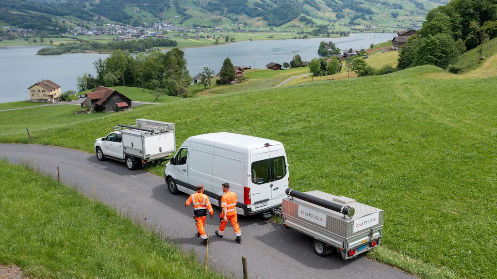 Zwei Cablex-Nutzfahrzeuge und Mitarbeitende in Warnkleidung auf einer Strasse in ländlicher Umgebung am See.