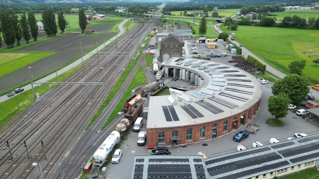 Luftaufnahme der historischen Rundbauremise und des Bahnhofsgebäudes Sargans mit schwarzen Photovoltaikmodulen auf dem Dach, umgeben von Gleisanlagen.