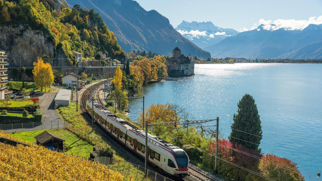Ein Personenzug fährt entlang eines Sees, umgeben von herbstlicher Landschaft und einer Bergkulisse.